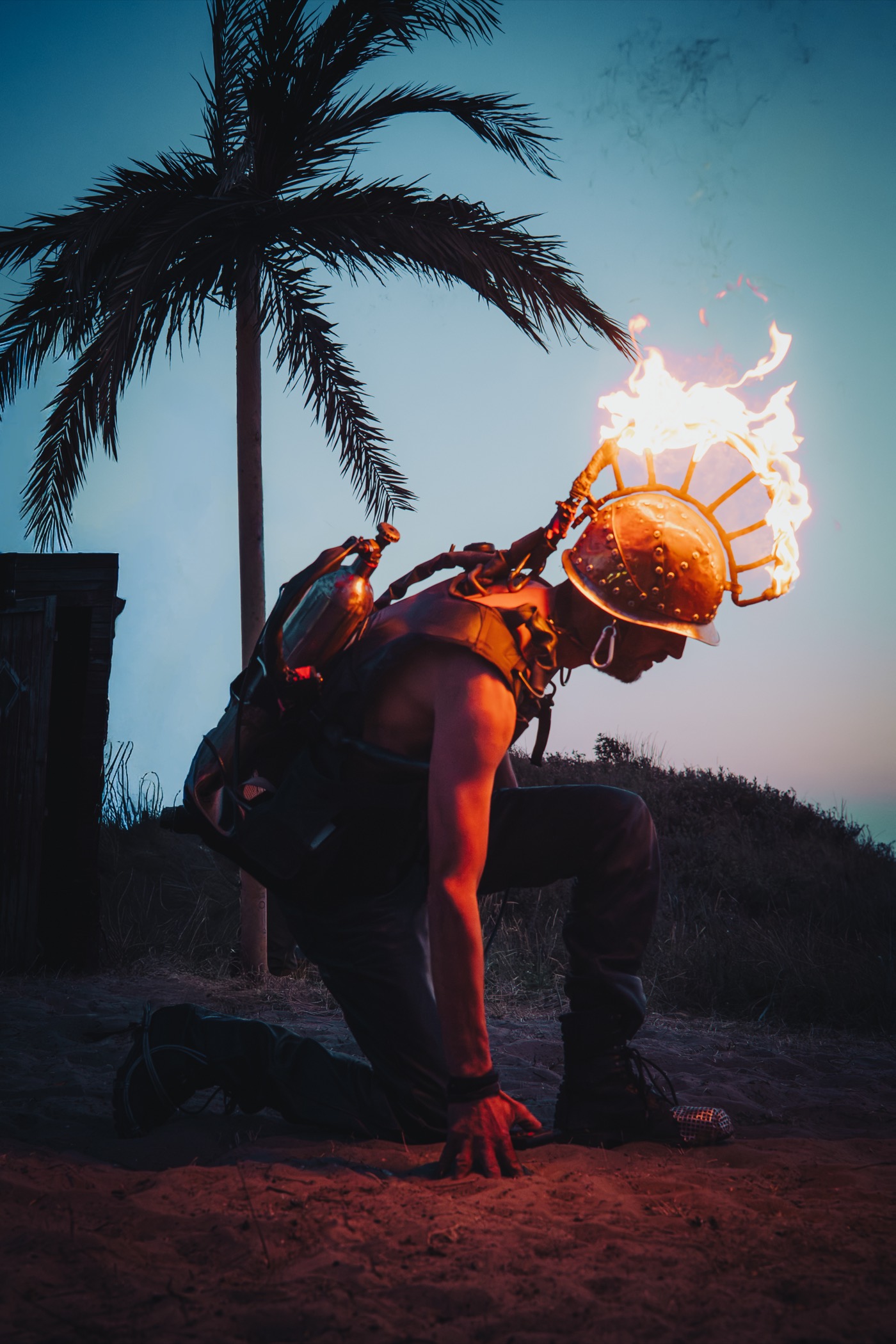 A fire performer in a flaming crown helmet kneeling under a palm at dusk, Africa Burn.